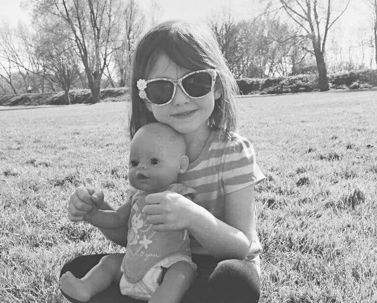 young girl wearing sunglasses, sitting on grass and holding dolly