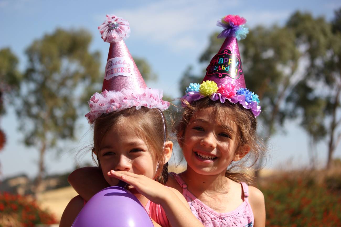 two young girls wearing party hats outside, and one of them blowing up a balloon