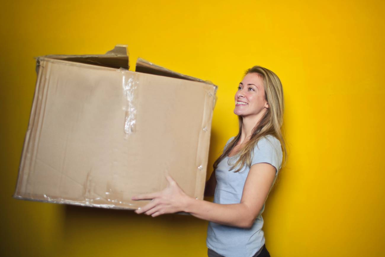 a blonde woman standing next to a bright yellow wall holding a cardboard box
