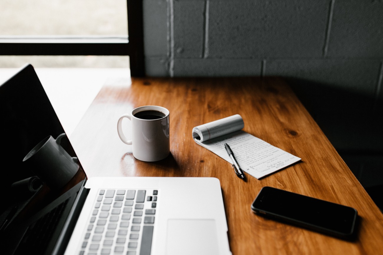 brown wooden desk with open laptop, mug, notepad, pen and phone