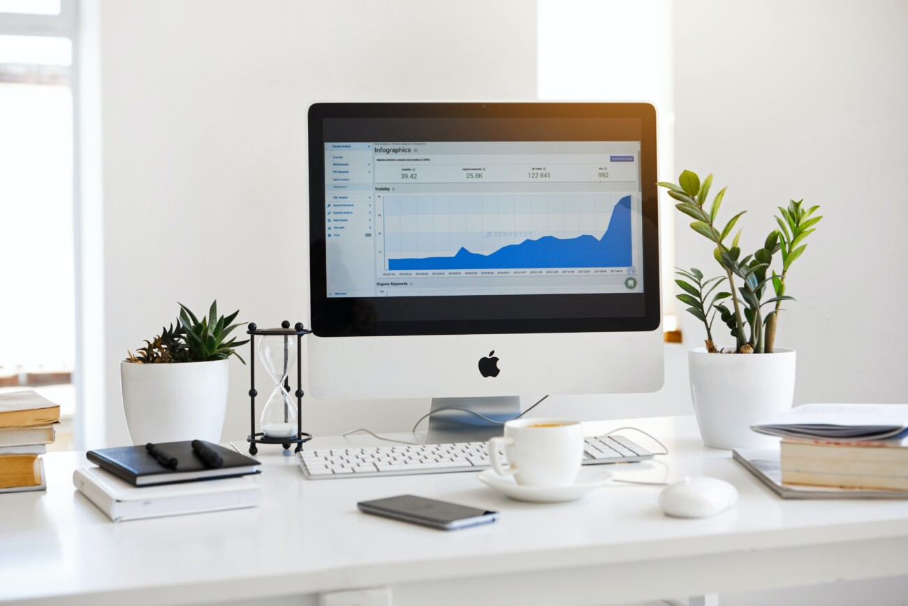 mac displaying blue graph on white desk with mug, notebook and plants