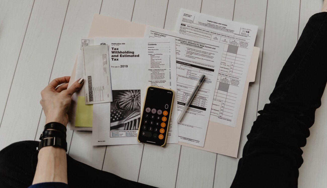 persons hand holding onto paper next to more piles of documents, calculator on phone and pen