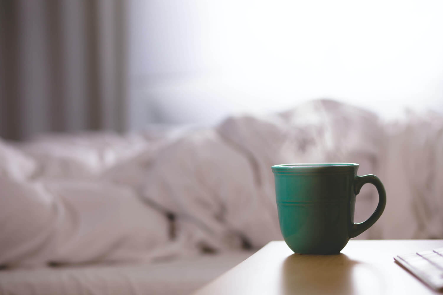 green mug with steam coming out of it on bedside table next to bed with white bedding bunched up