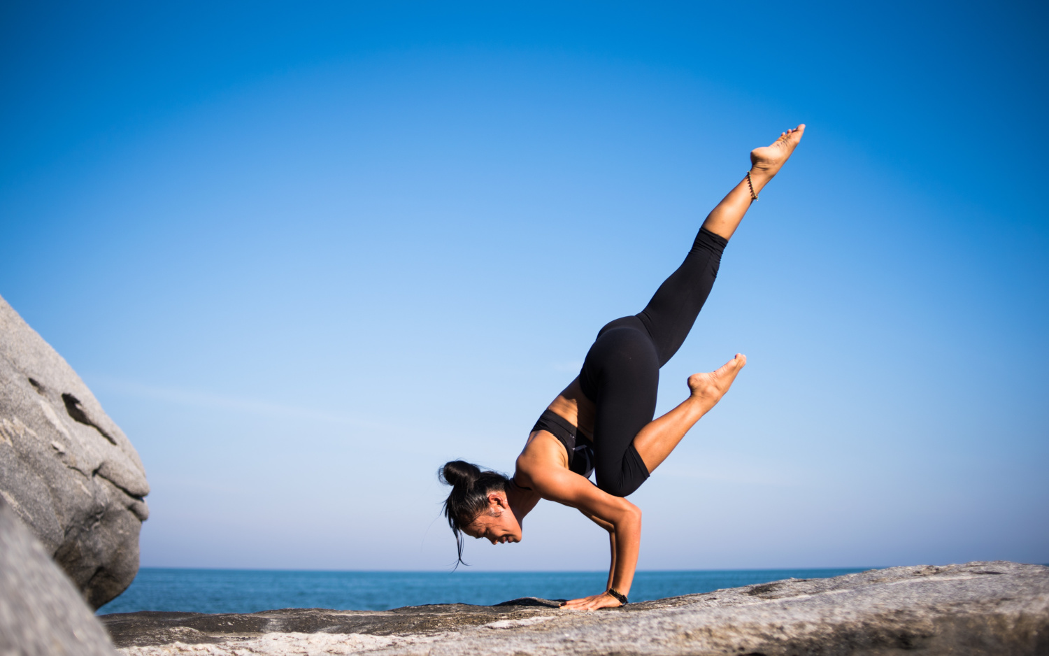 woman doing a yoga handstand pose on a rock with blue sky and sea behind her