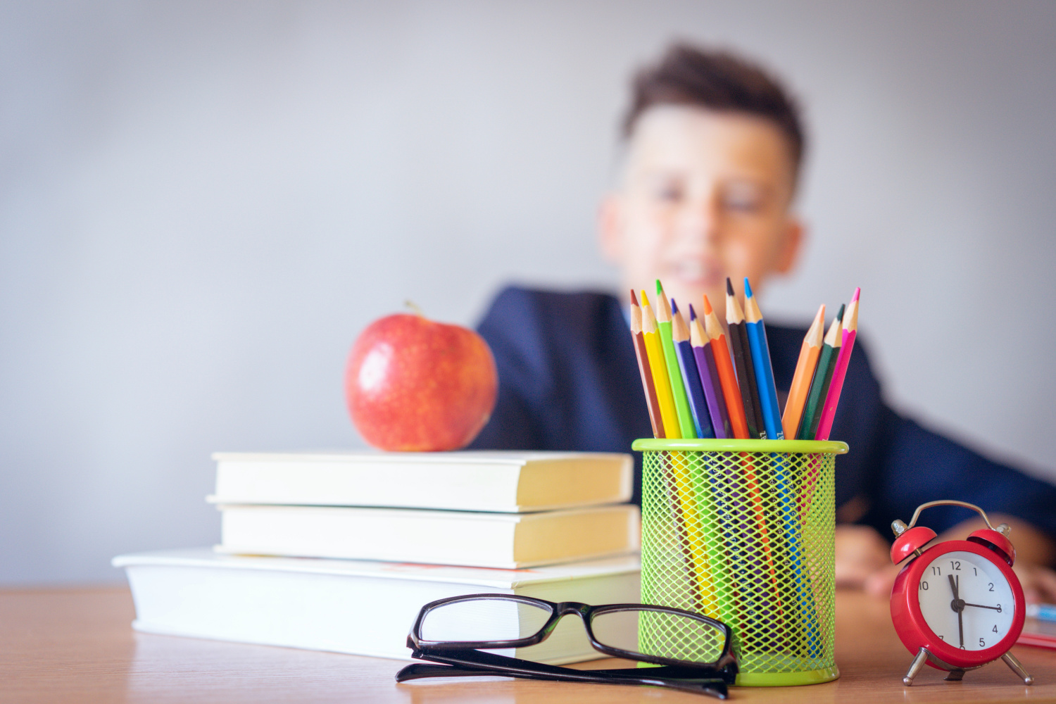 boy sitting at desk with a pile of books with an apple on top and glasses and a pot of crayons