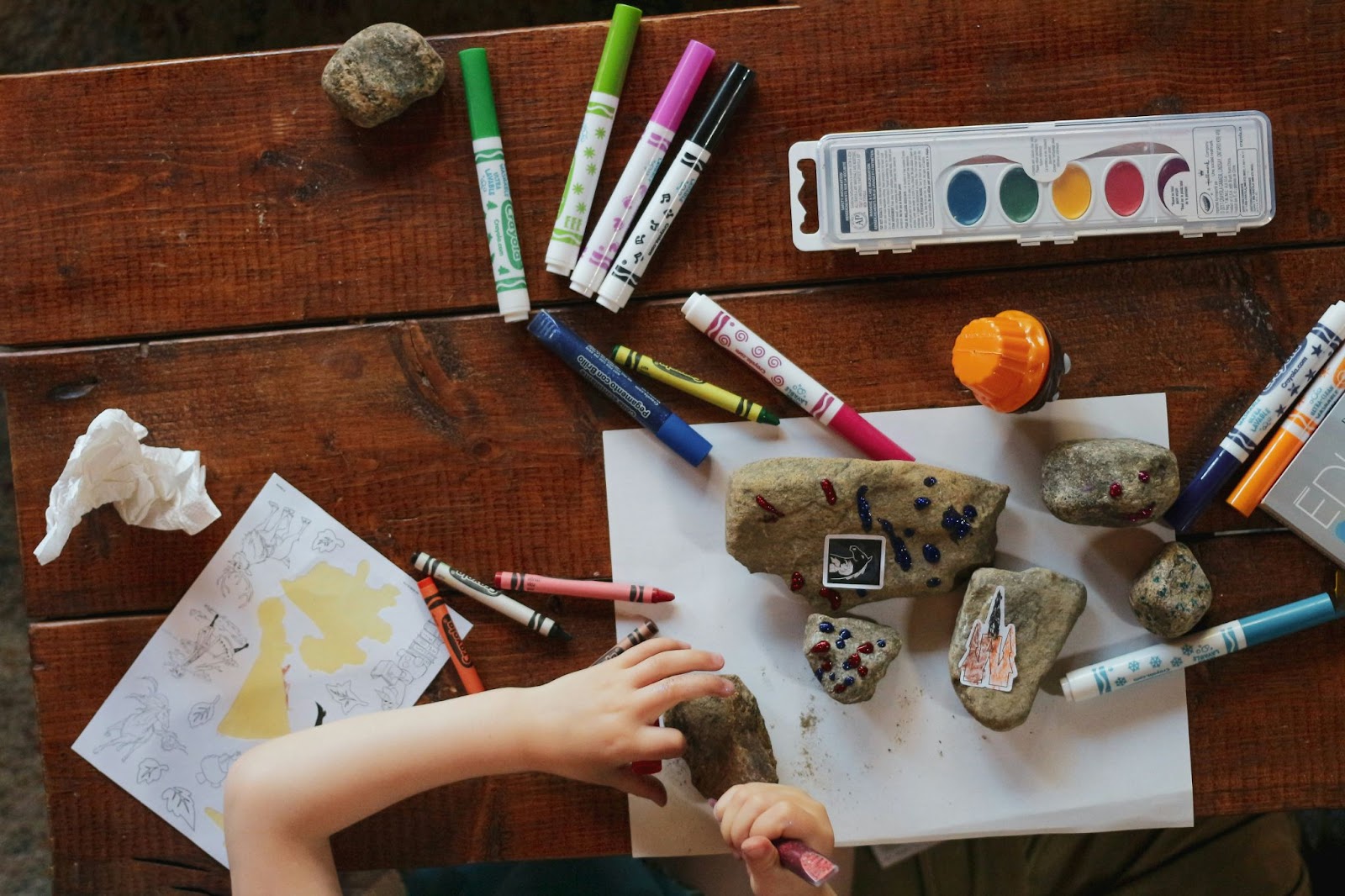 table from above filled with felt tips and paint and paper and child's hands