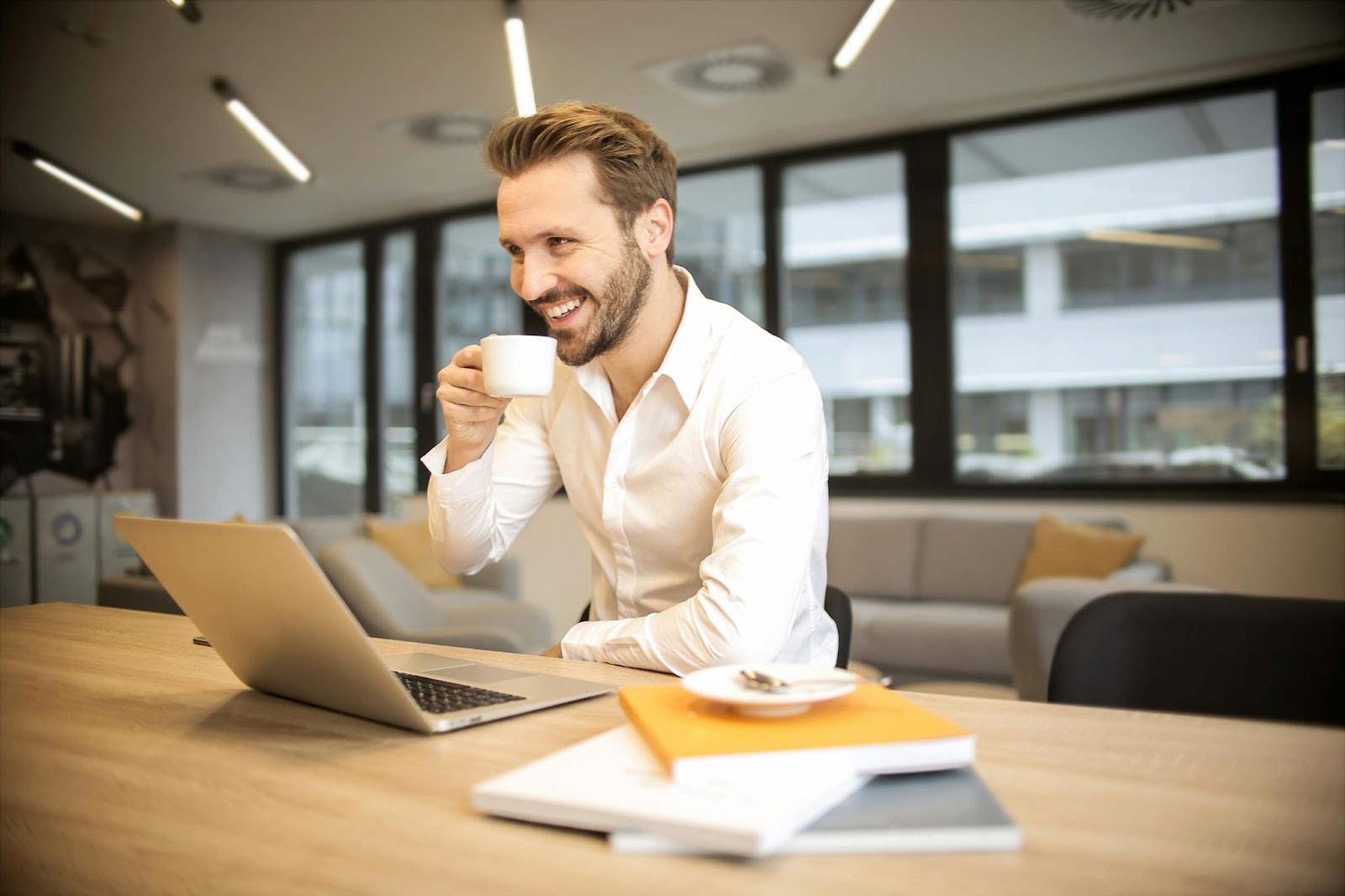 man sat at desk in office with laptop in front of him and holding a coffee cup and smiling