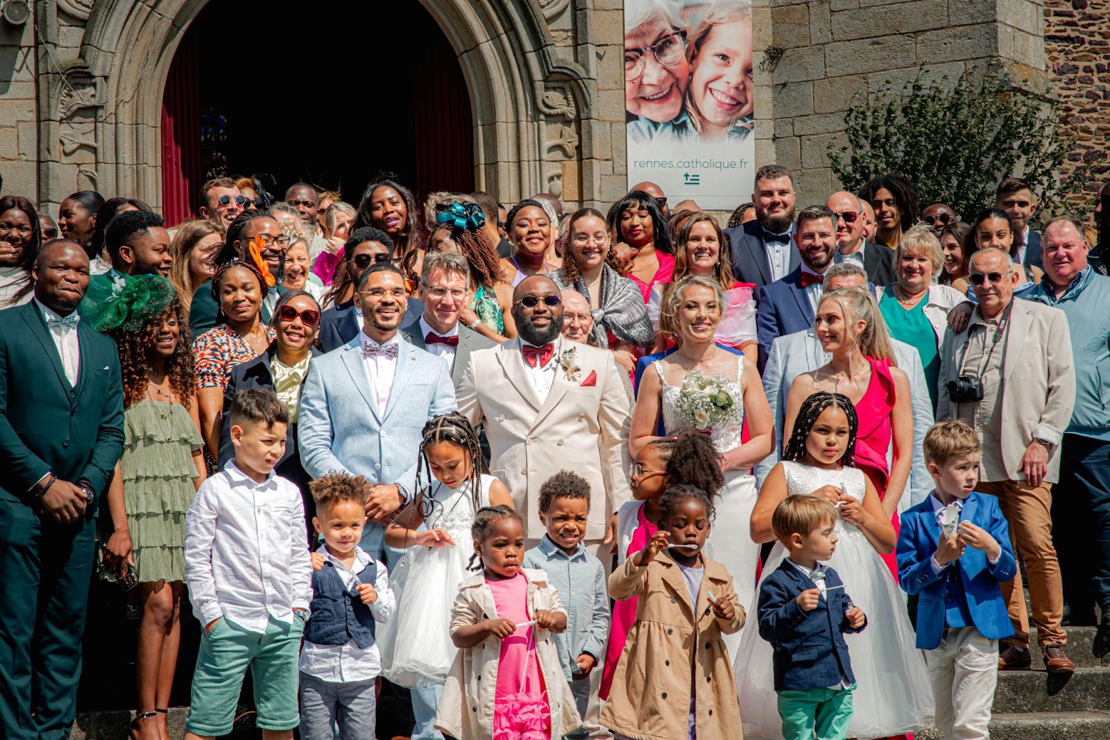 Wedding party with bride and groom outside church. People look happy and lots are wearing bright colours.