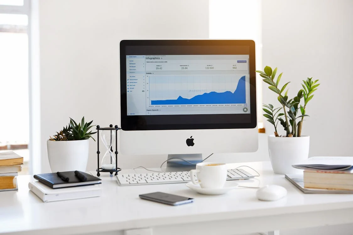 mac computer on white desk with plants, notebook and coffee cup