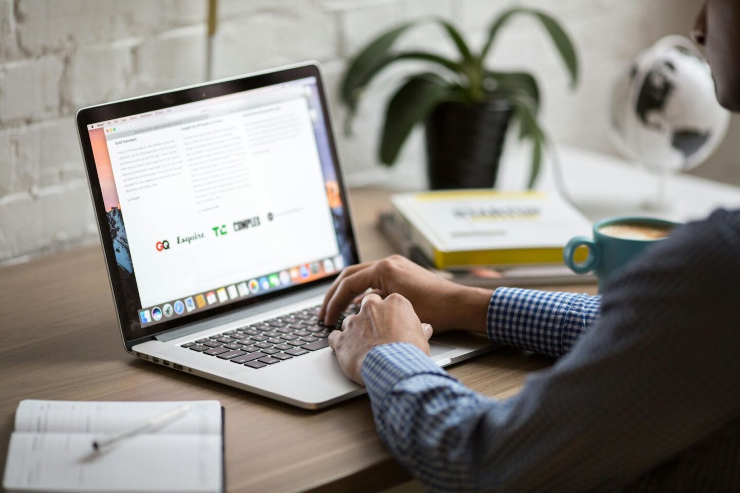 person wearing checked shirt with hands on laptop on brown desk next to plant and coffee cup
