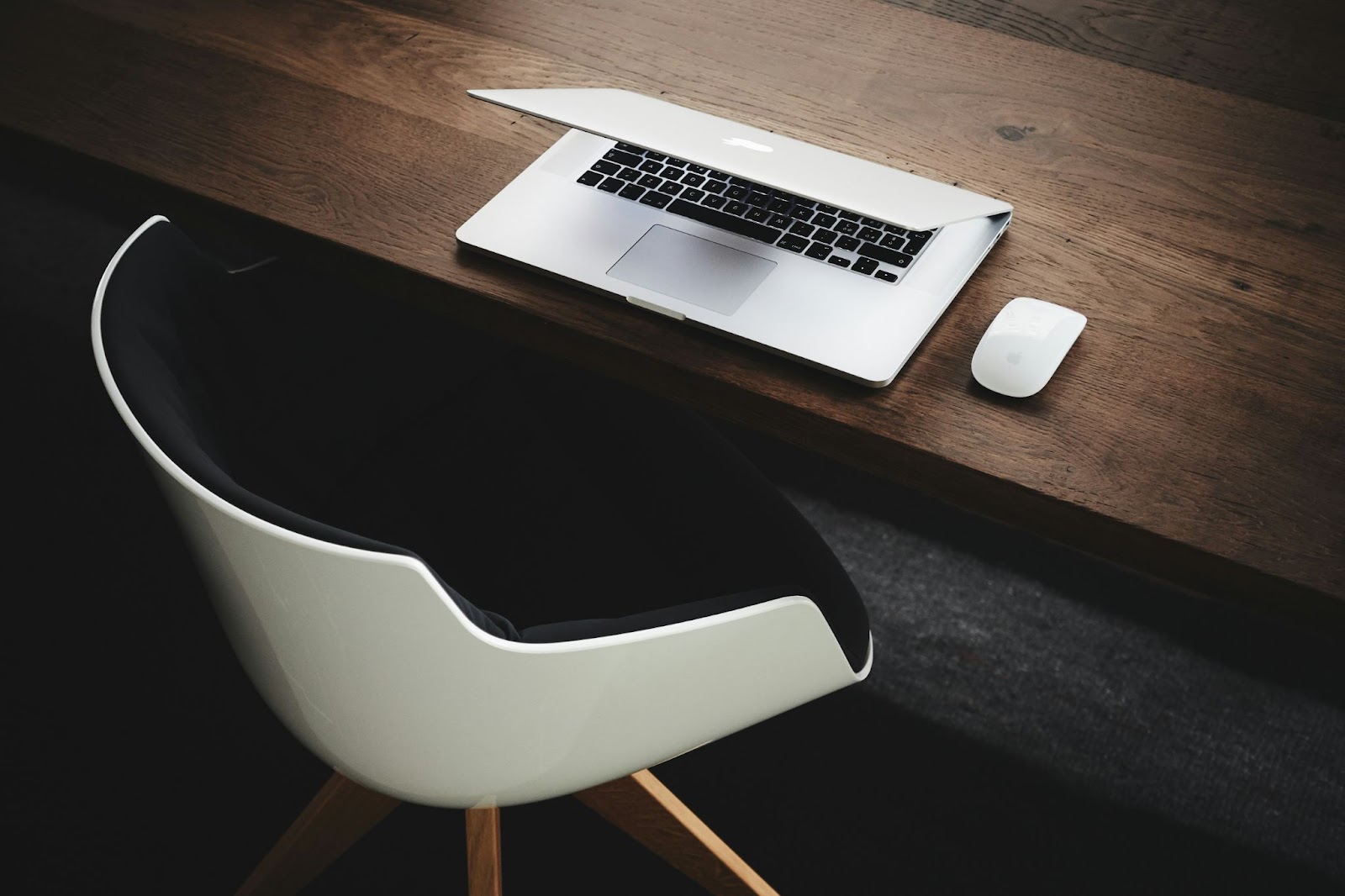 brown wooden desk with a partially closed laptop and white mouse on it and a white and black chair next to it.
