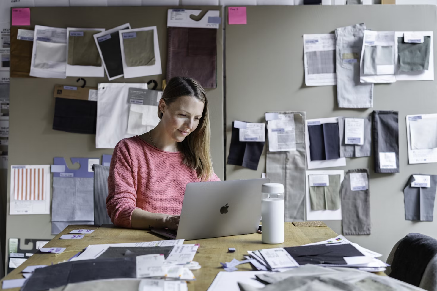 woman in pink top sitting at desk at laptop with lots of papers on desk and pinned on wall