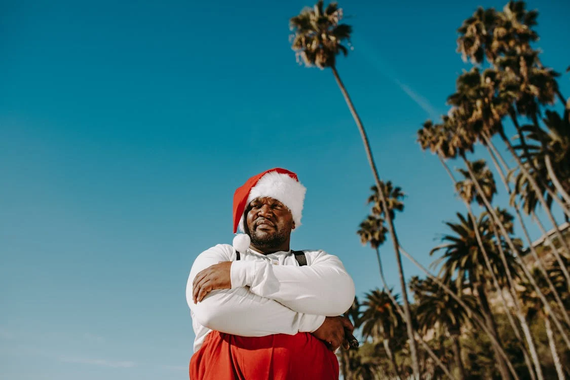 man dressed as santa with arms folded, blue sky and palm trees behind him