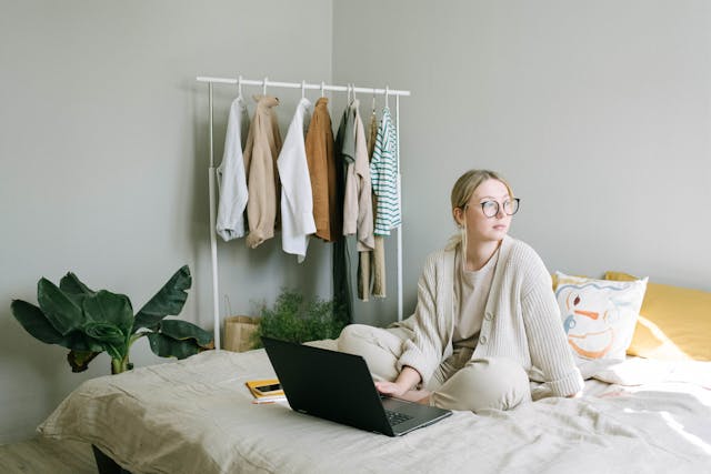 woman sitting on ded with hand on laptop, clothes in muted tones hanging on rail in background