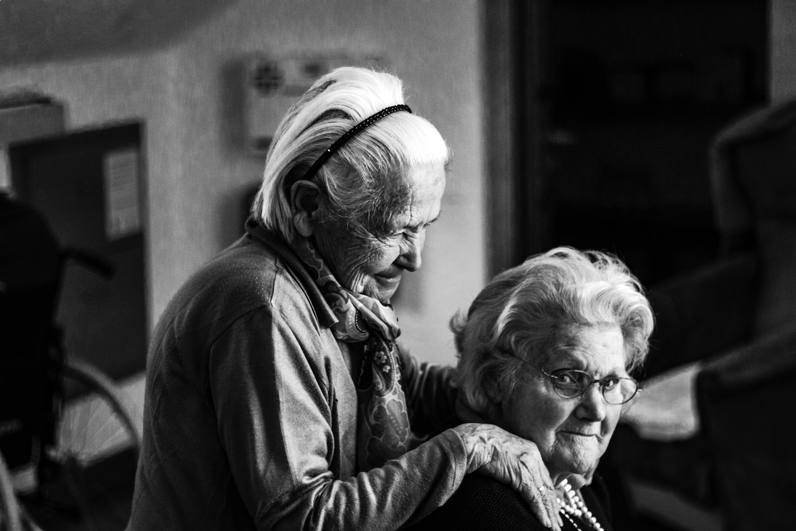 greyscale photo of older woman standing behind another older woman sitting on chair