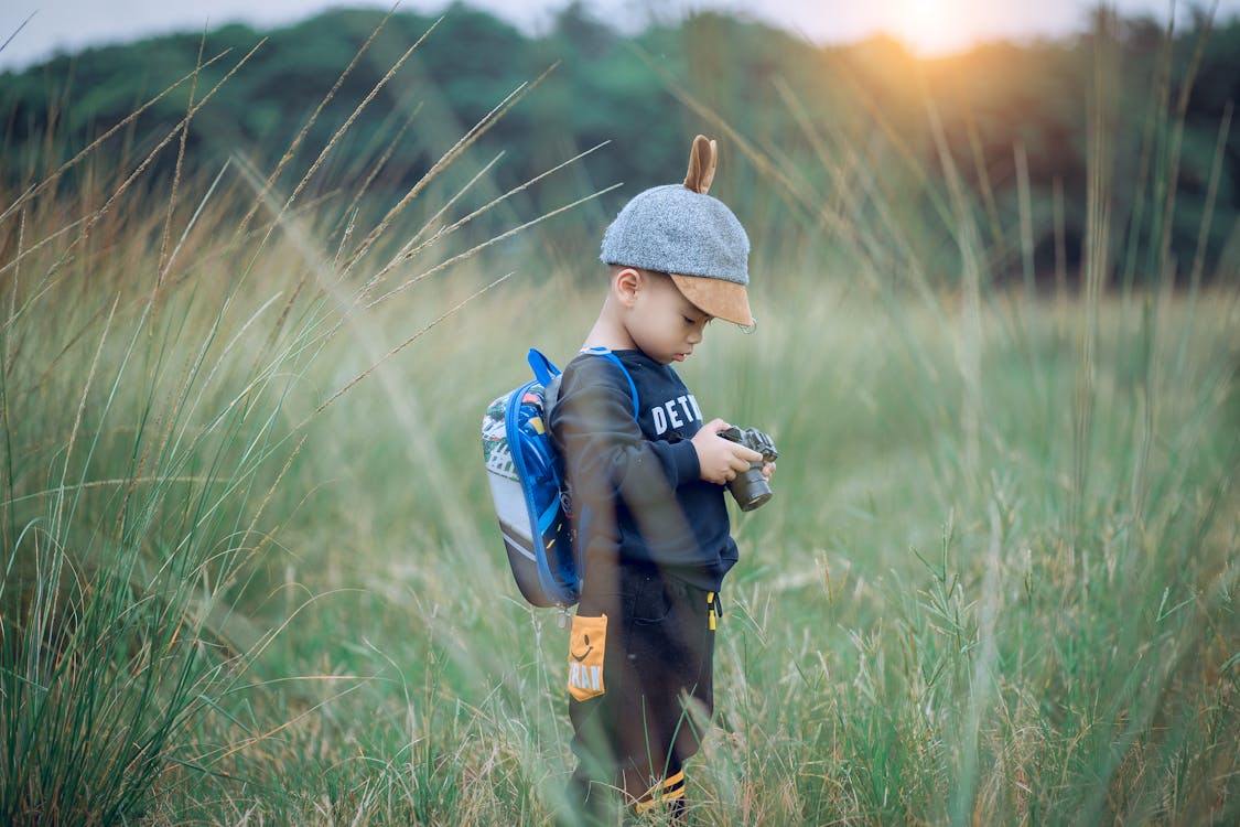 child wearing backpack, standing in long grass looking at camera in hand