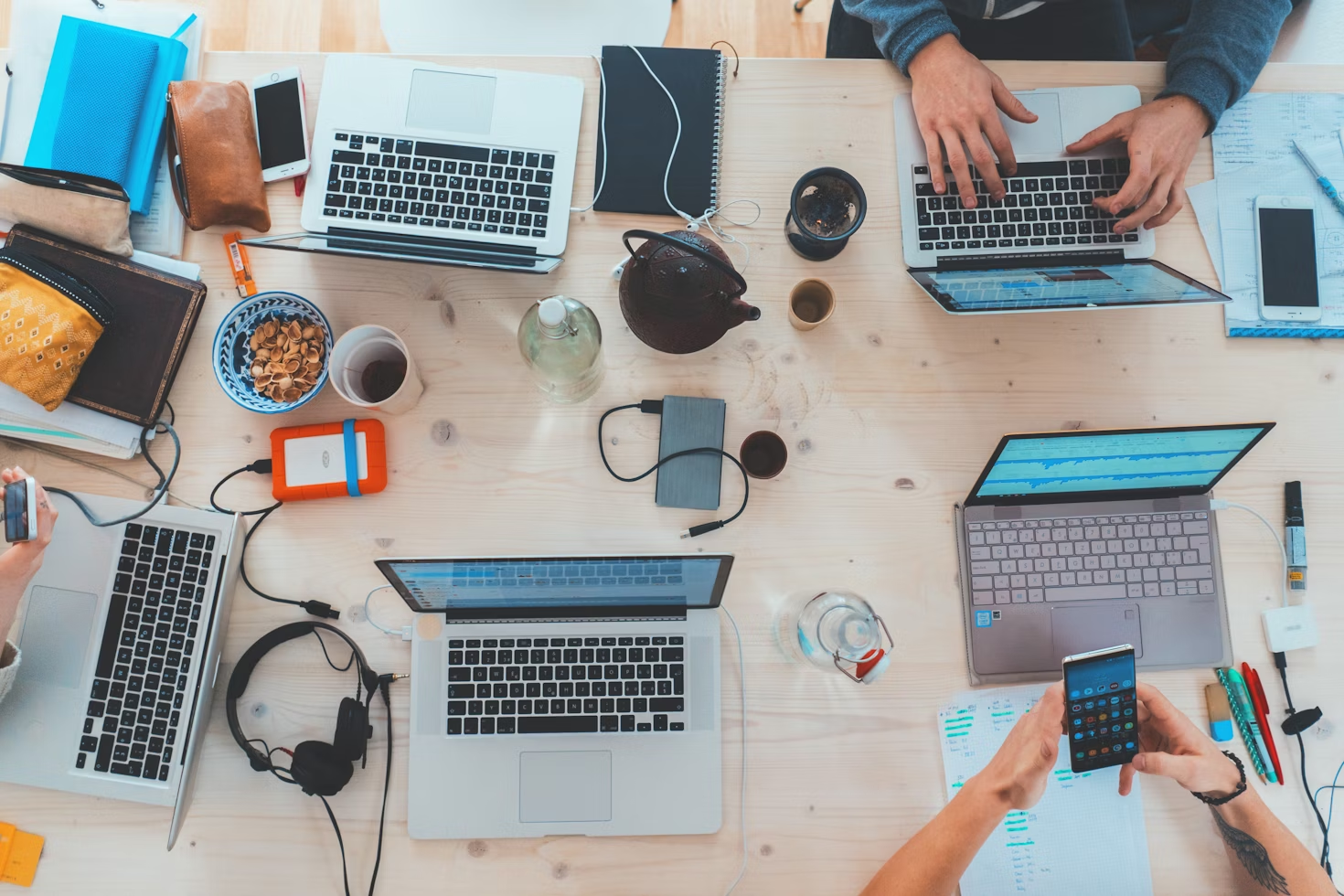 view of large desk from above with laptops, cups of coffee, phones, chargers and wires with people working on laptops