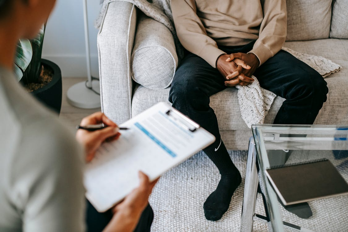 bottom half of person sitting on sofa in room having an appointment with another person with form on clipboard in their hands