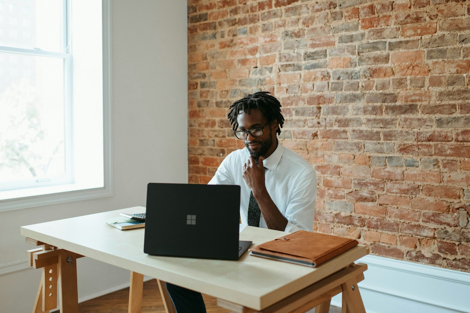 man sitting at a desk with laptio open and a brick wall behind him