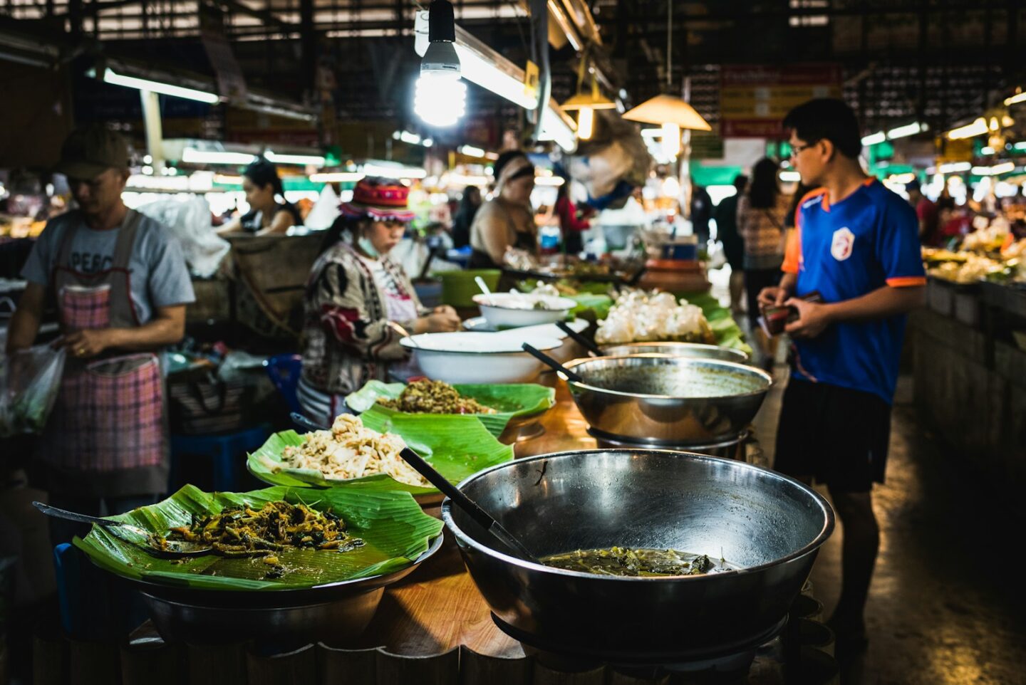 man in blue t-shirt standing in front of people making asian food at outdoor market a