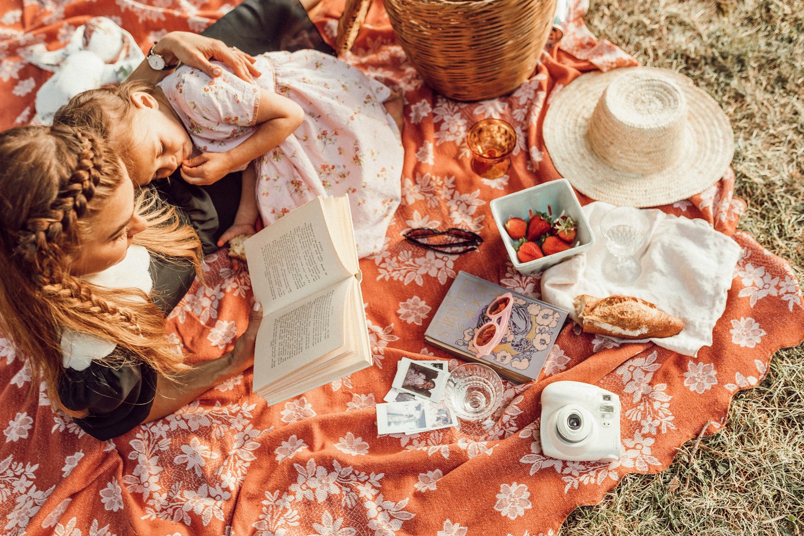 woman reading with girl while lying on orange and white floral picnic blanket on grass. With strawberries in bowl, a hat, polaroid camera, basket, glasses