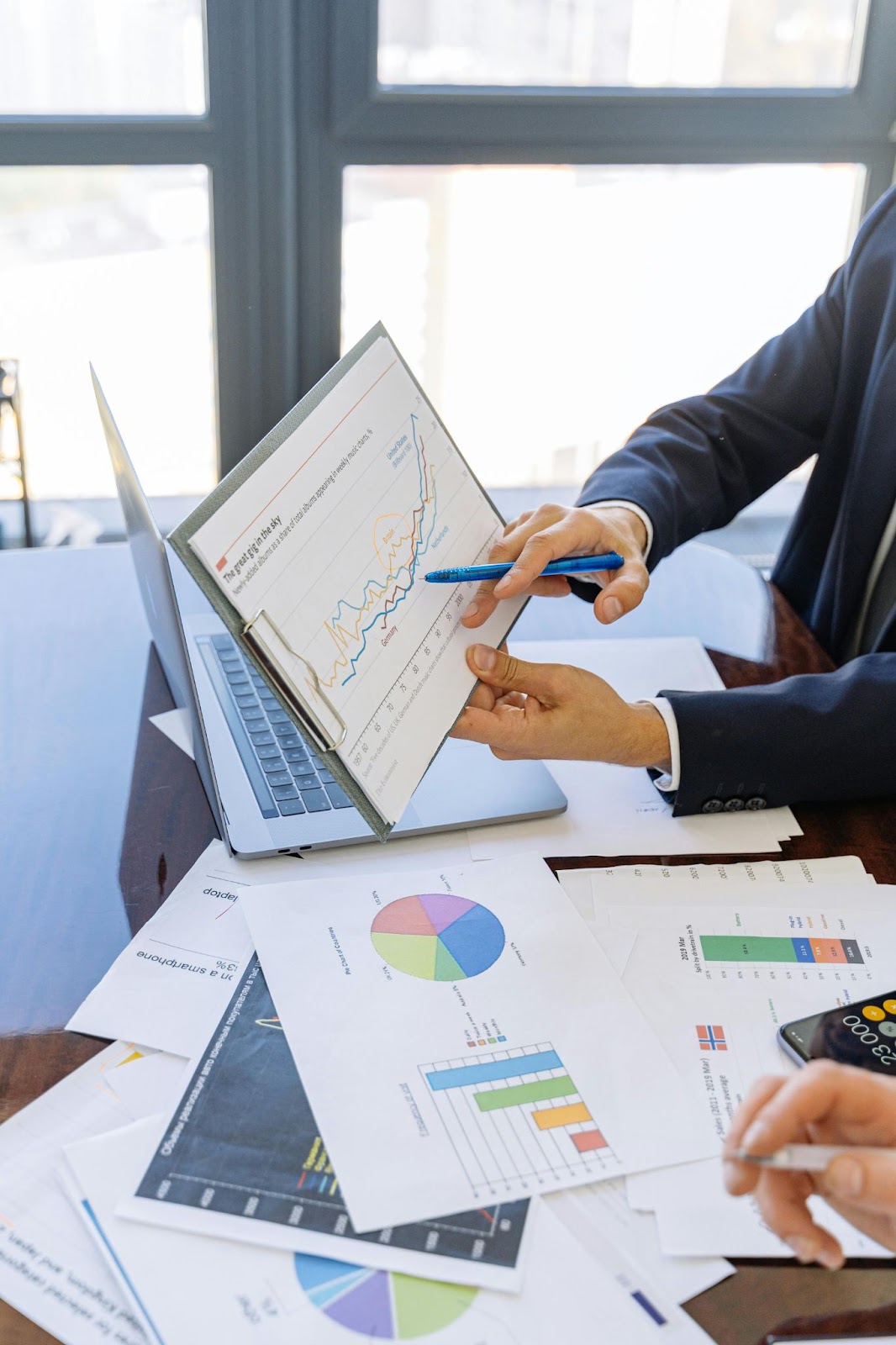man in suit holding pen pointing at chart, with more charts on desk and laptop