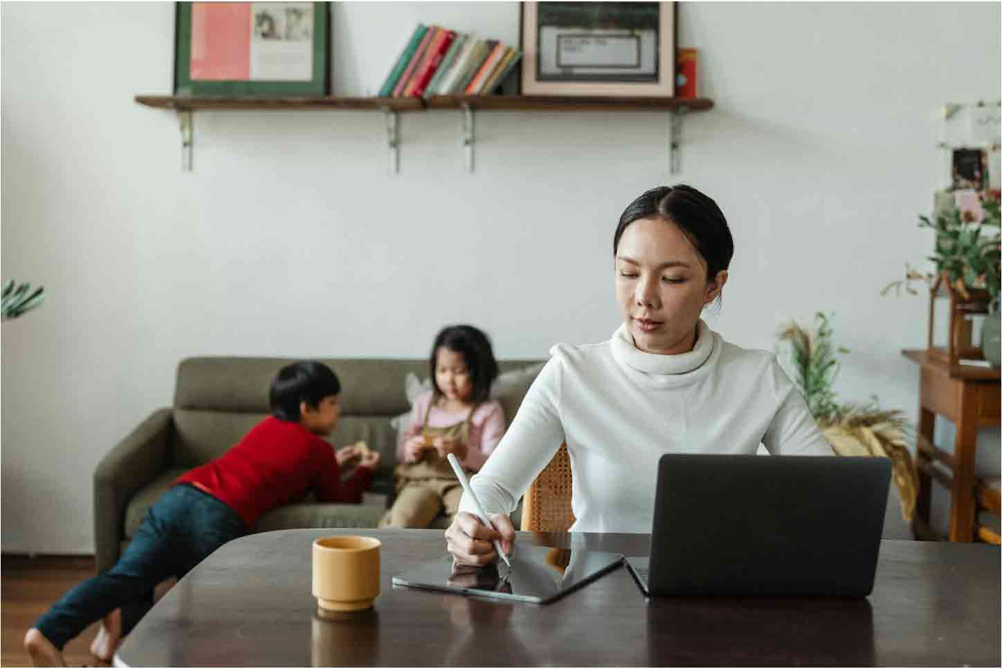 mum working at table at home whilst kids play in the background on grey sofa