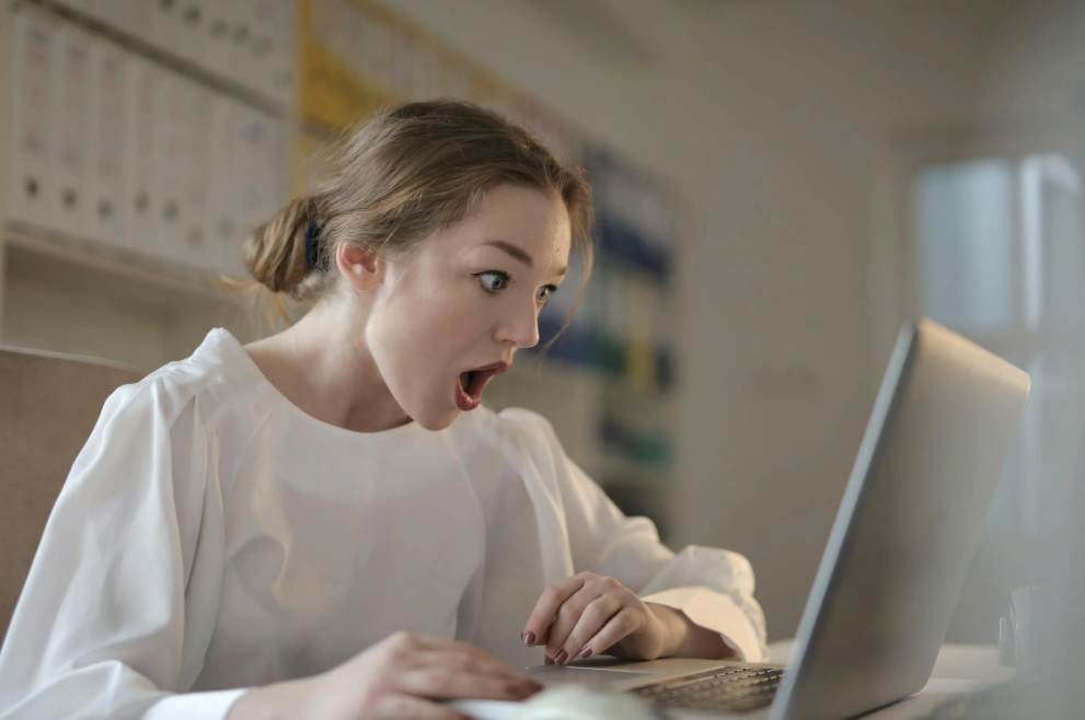 woman in white top sitting at laptop with her mouth open wide in shock