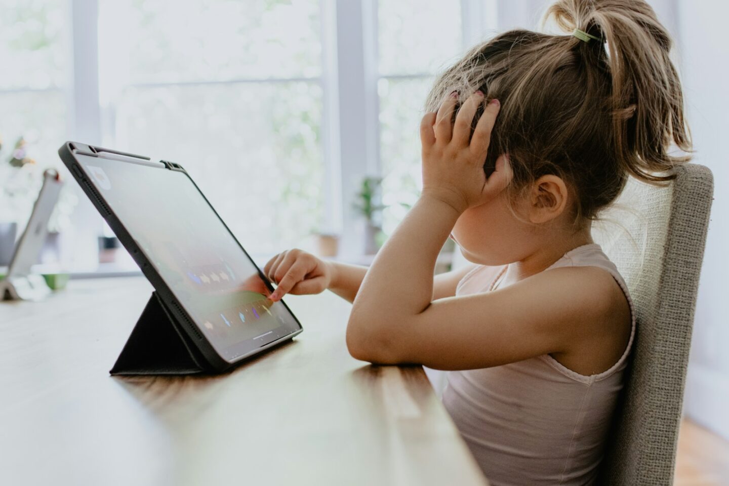 girl in white tank top using black tablet computer at table with her hand resting on her head
