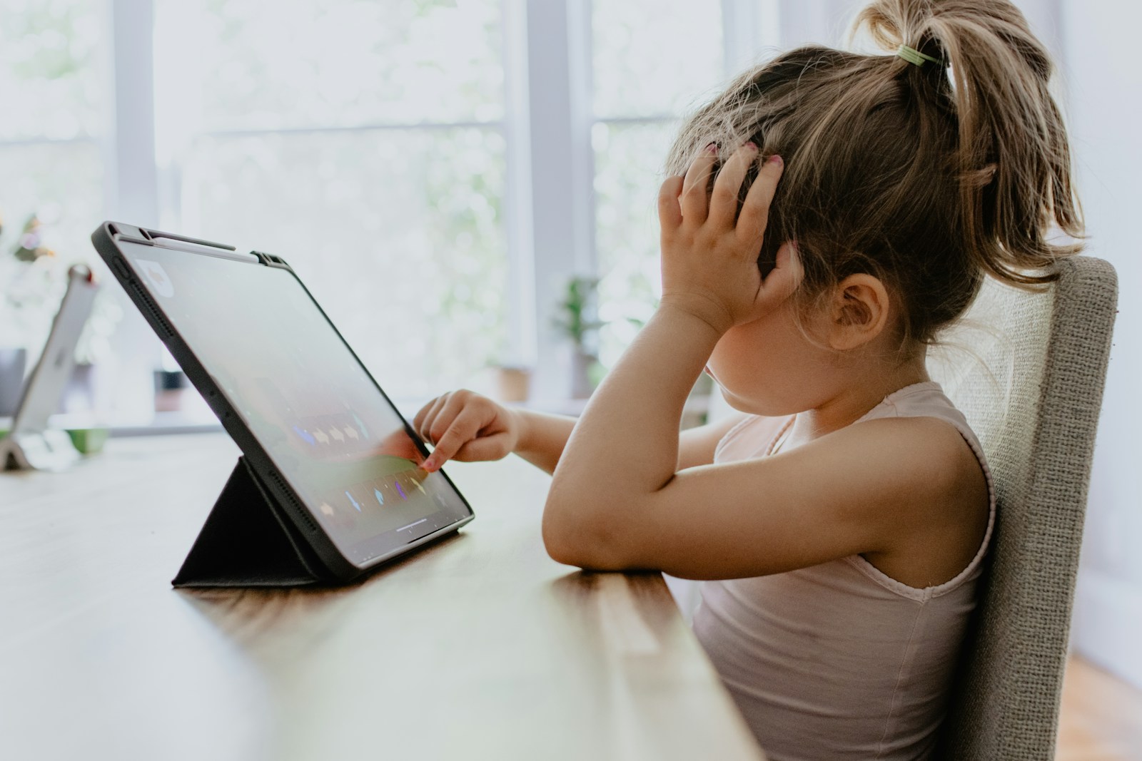 girl in white tank top using black tablet computer at table with her hand resting on her head
