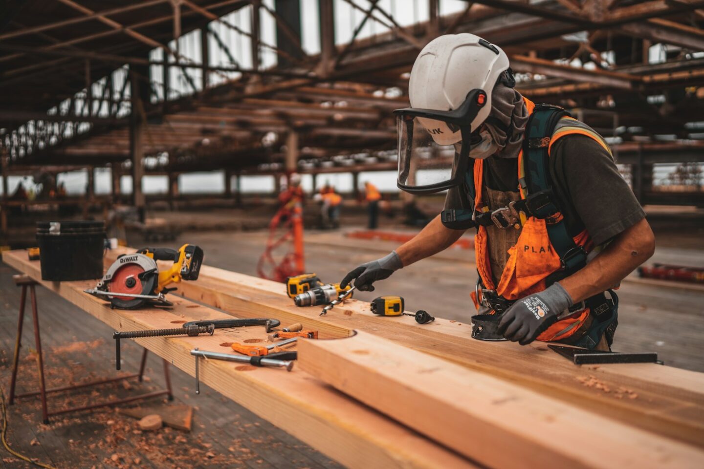 man in orange and black vest wearing white helmet holding yellow and black power tool and working on planks of wood