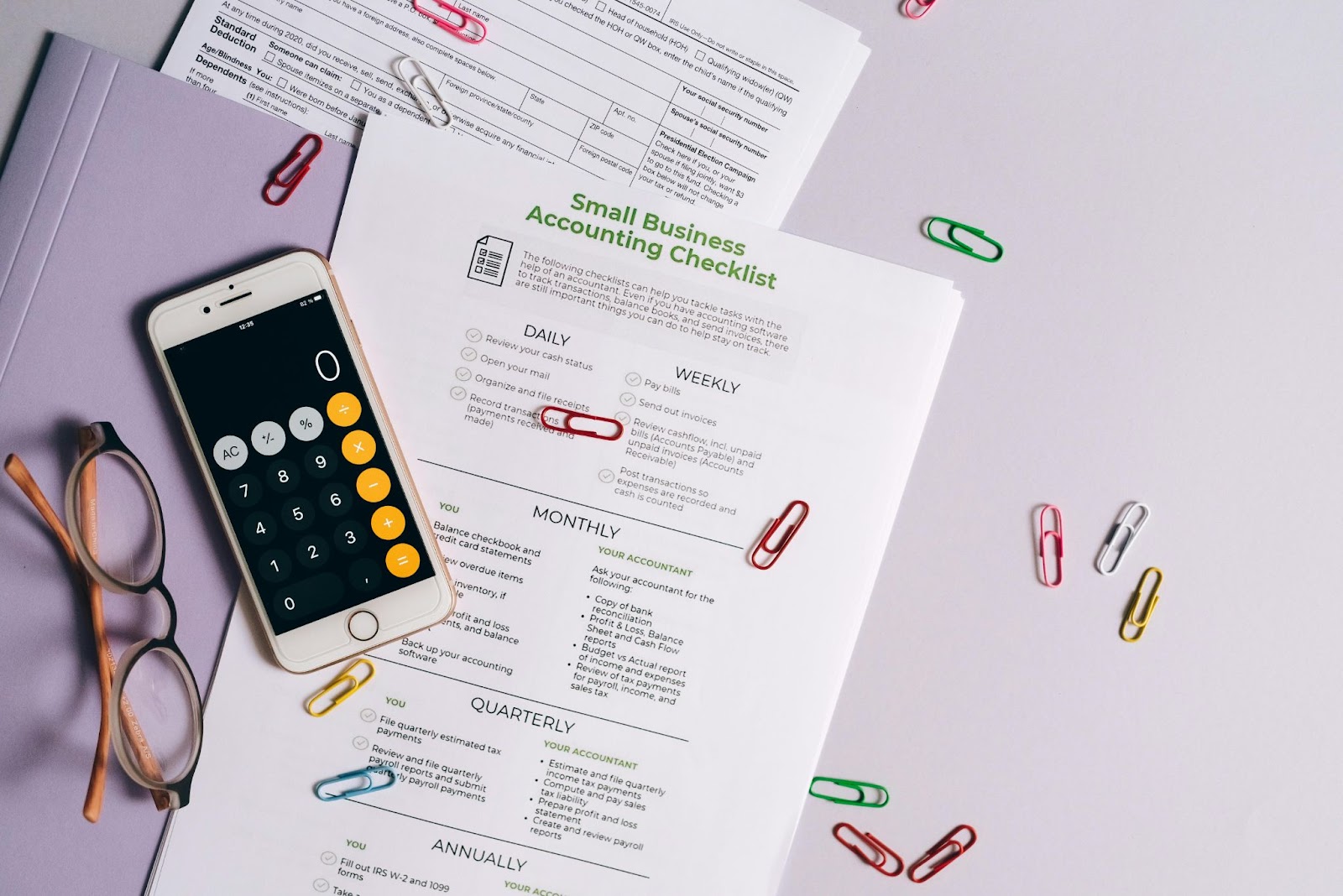 paper with small business accounting checklist on, and scattered paper clips, phone with calculator on screen, and glasses