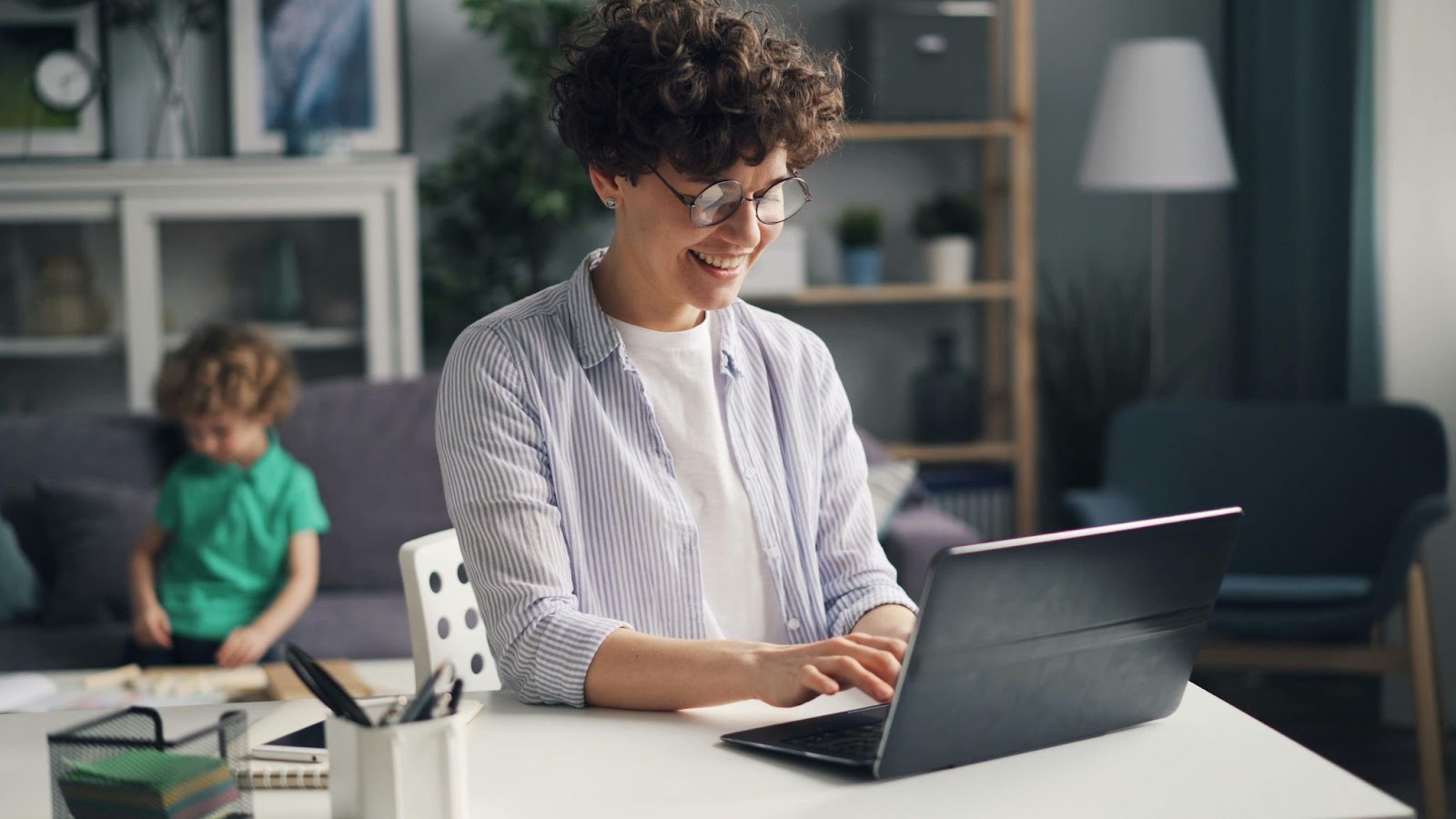 woman with short curly hair sitting at laptop and smiling, with toddler in the background