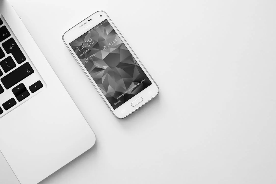 black and white photo of white phone next to laptop