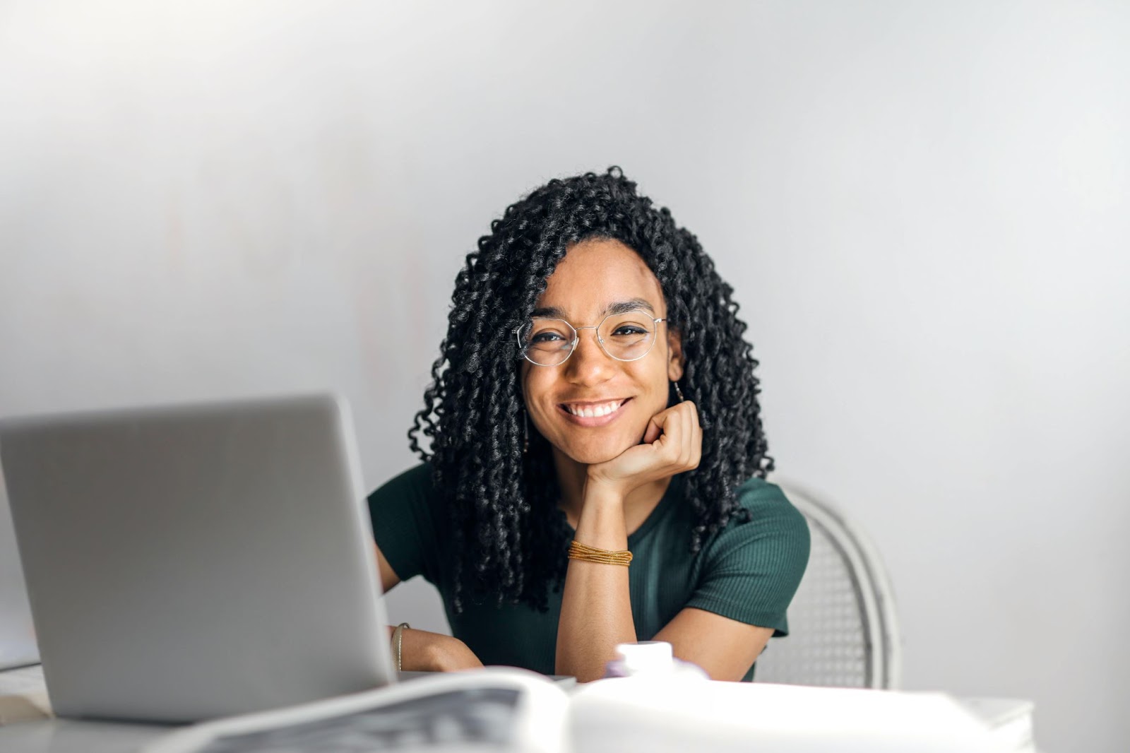 woman with curly hair sitting at desk with laptop in front of her, smiling and resting her head on her hand