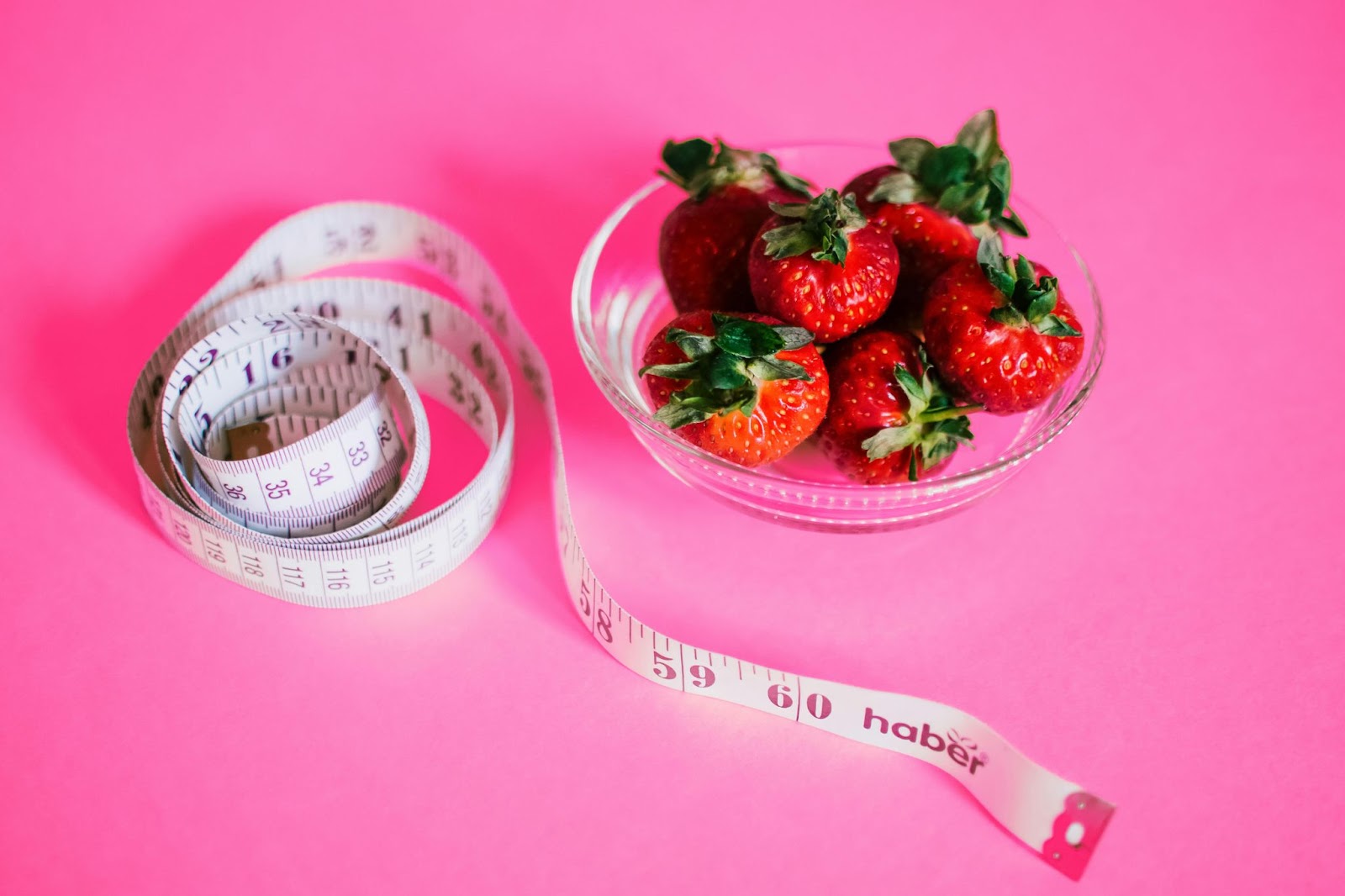 strawberries in glass bowl next to tape measure with bright pink background