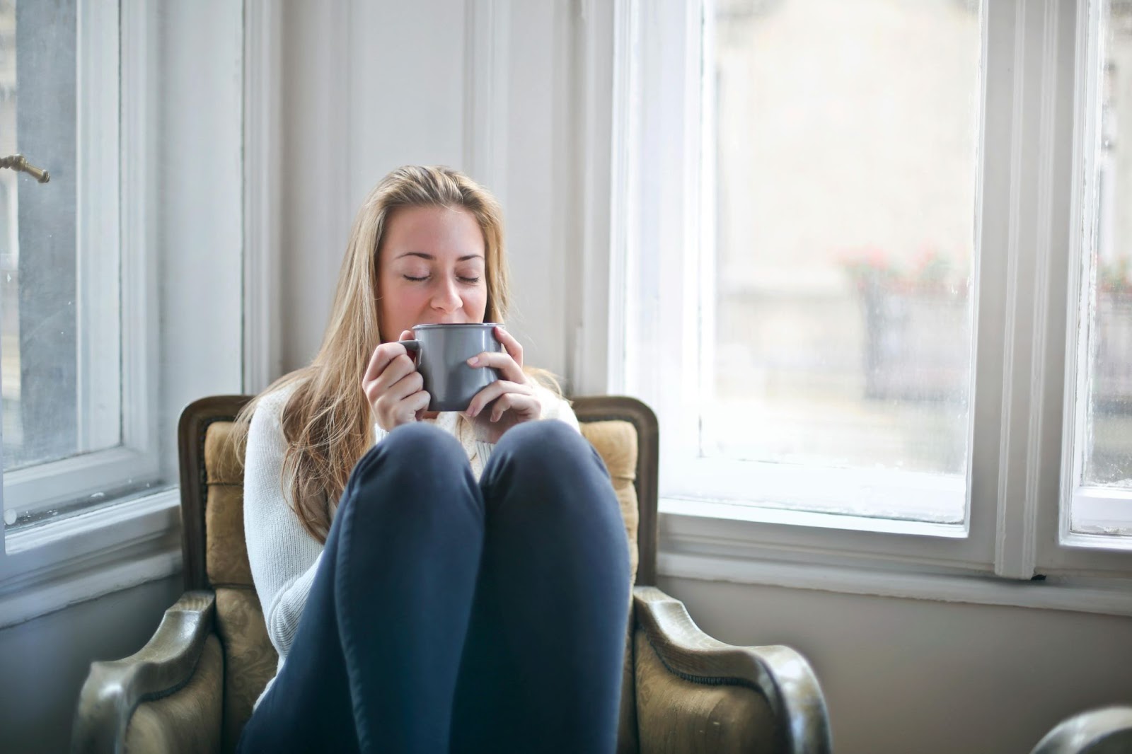 woman sitting on chair with her knees up and eyes closed, enjoying a hot drink in a blue mug