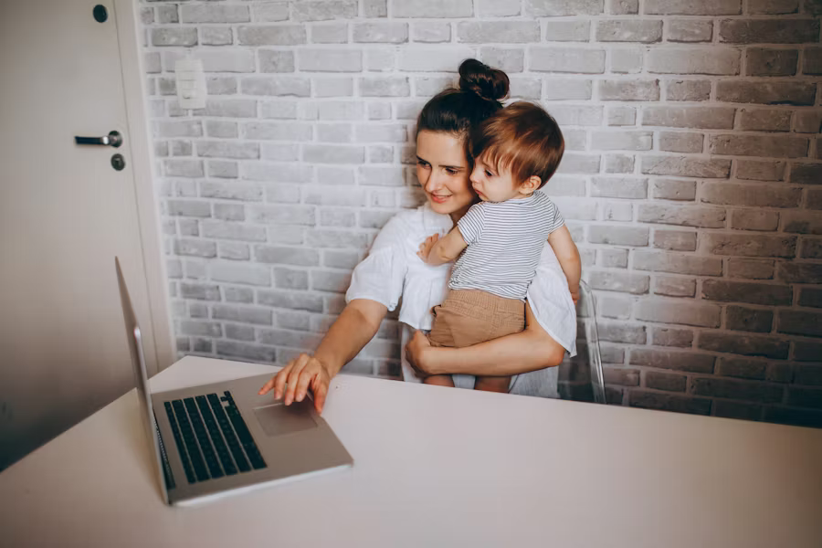 mother sitting at table on laptop and holding child in arms