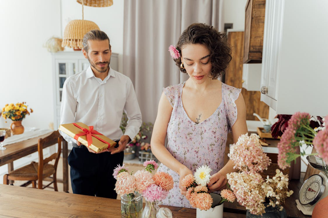 man walking towards woman arranging flowers with wrapped gift in his hand