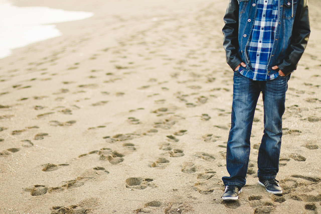 person on sandy beach wearing blue jeans, denim jacket, checked shirt and trainers