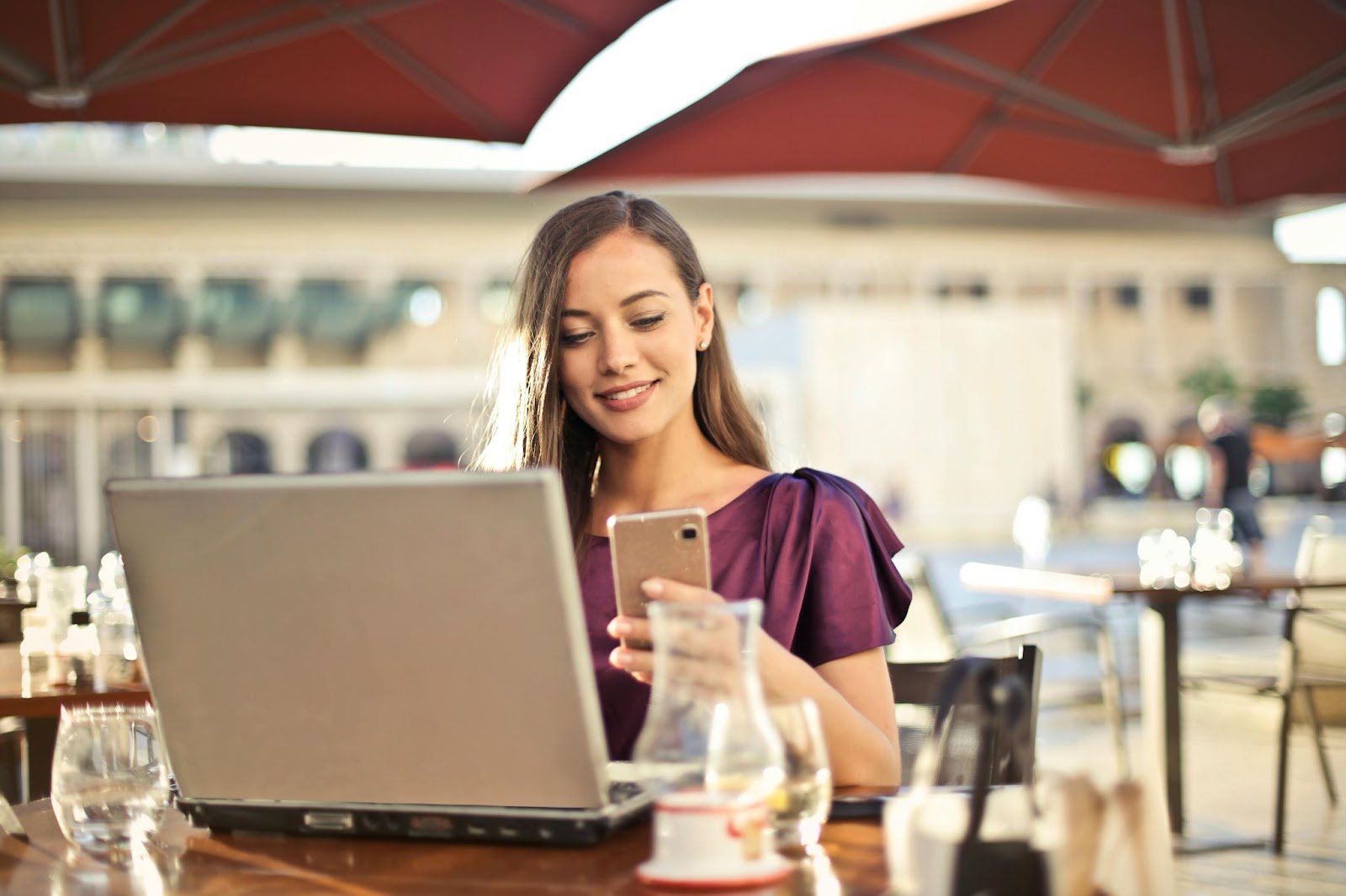 woman sitting at outdoor seating at cafe, with laptop in front of her and holding phone and smiling