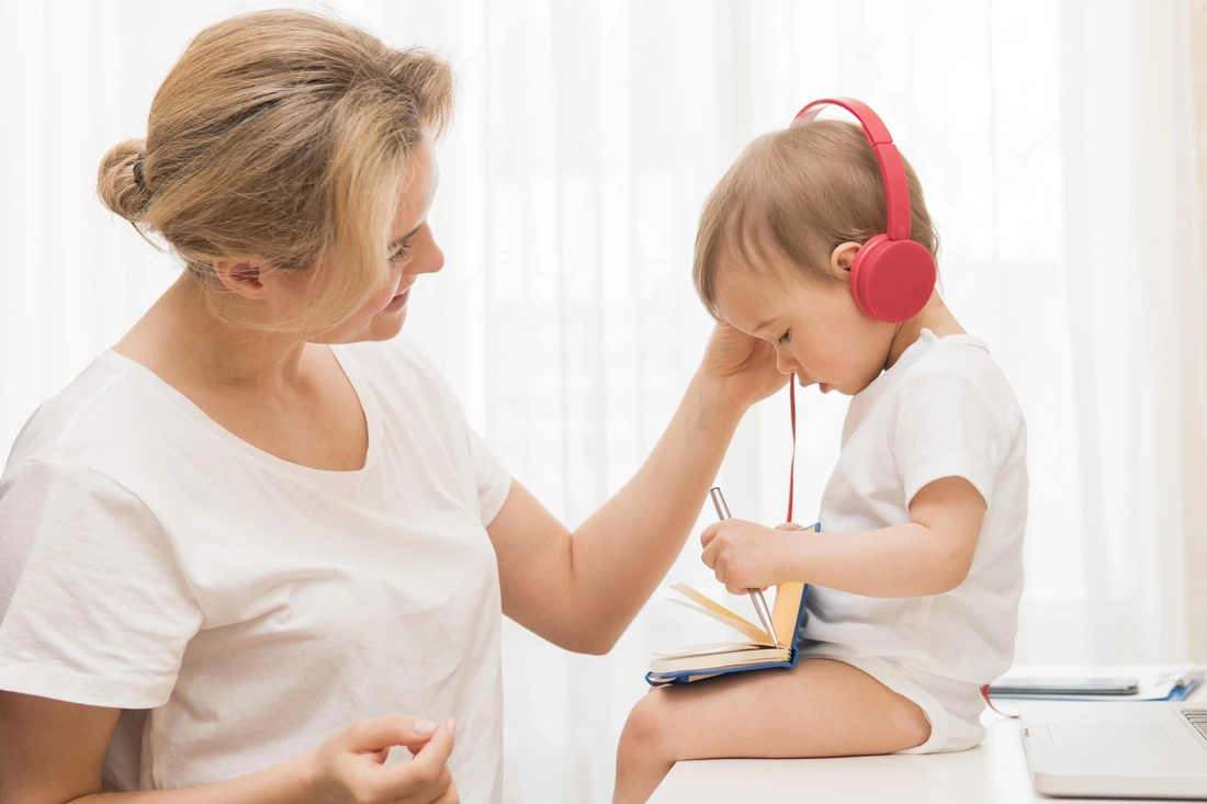 woman standing next to small child sitting on surface wearing red headphones, woman is smiling and has hand on childs ear