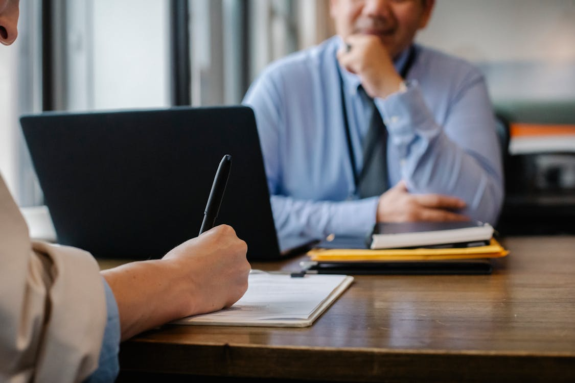 two men sitting at desk opposite each other, with laptops and pens and paper