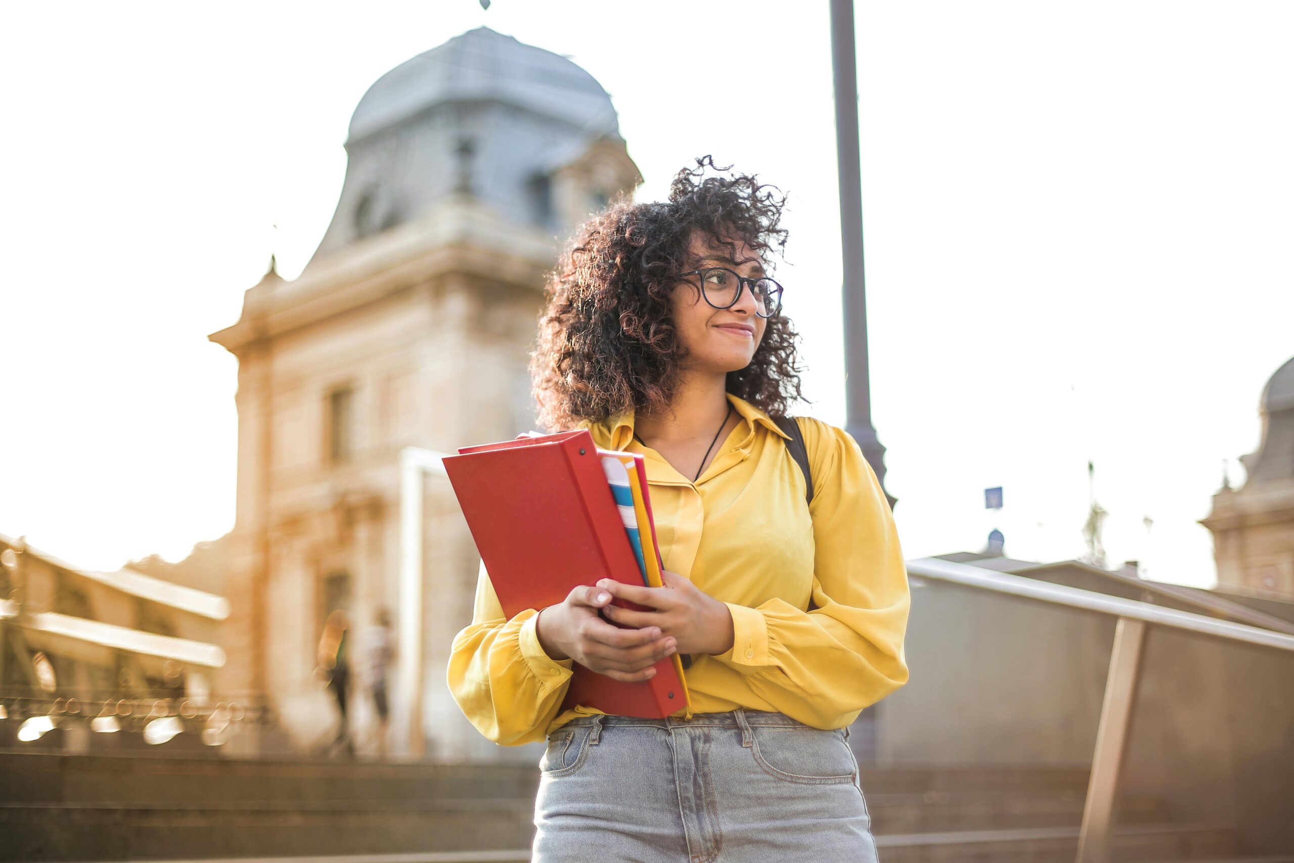 woman in yellow jacket and jeans, standing outside with books in her arms and smiling