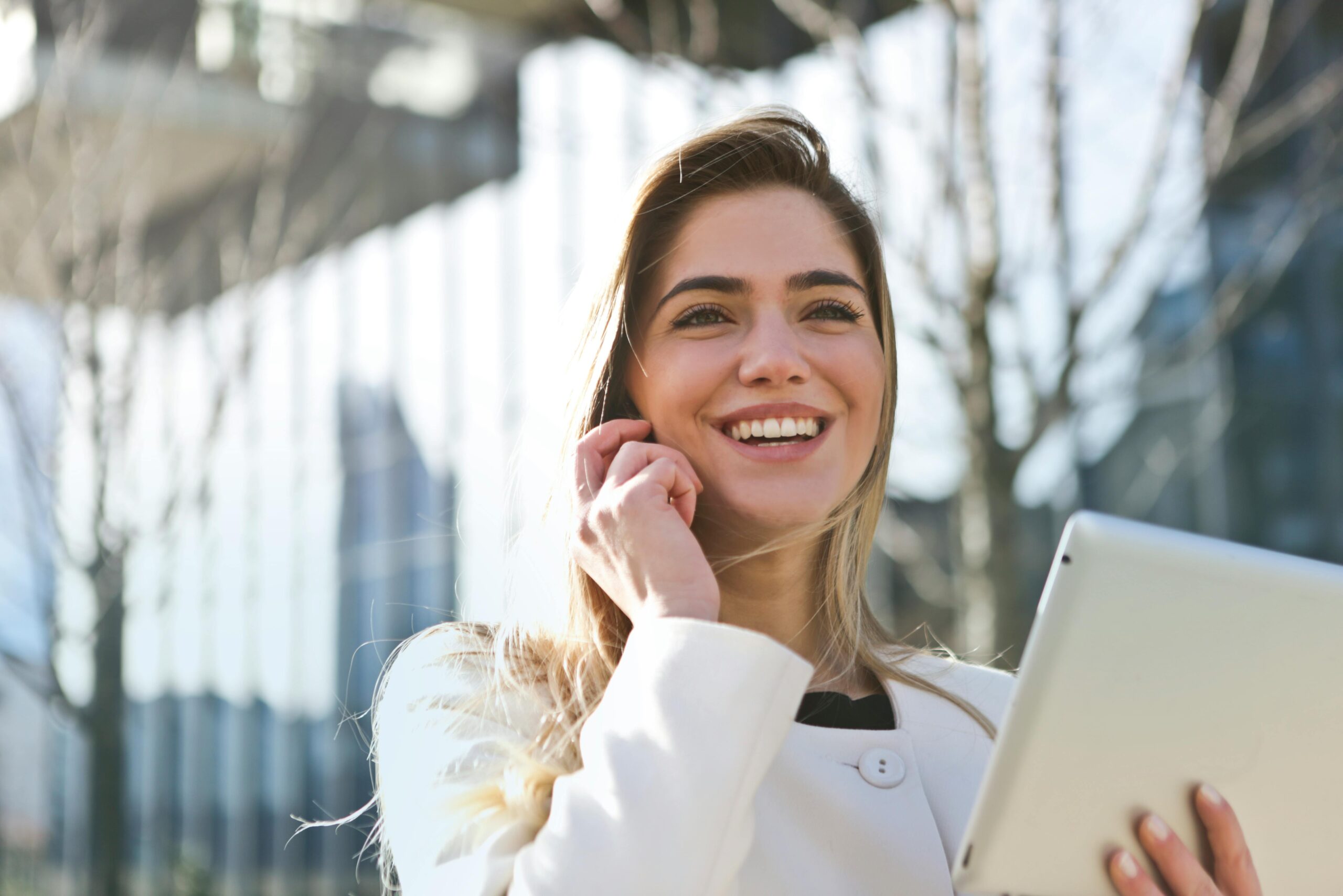 Woman outside, wearing white jacket on the phone, smiling, and holding iPad