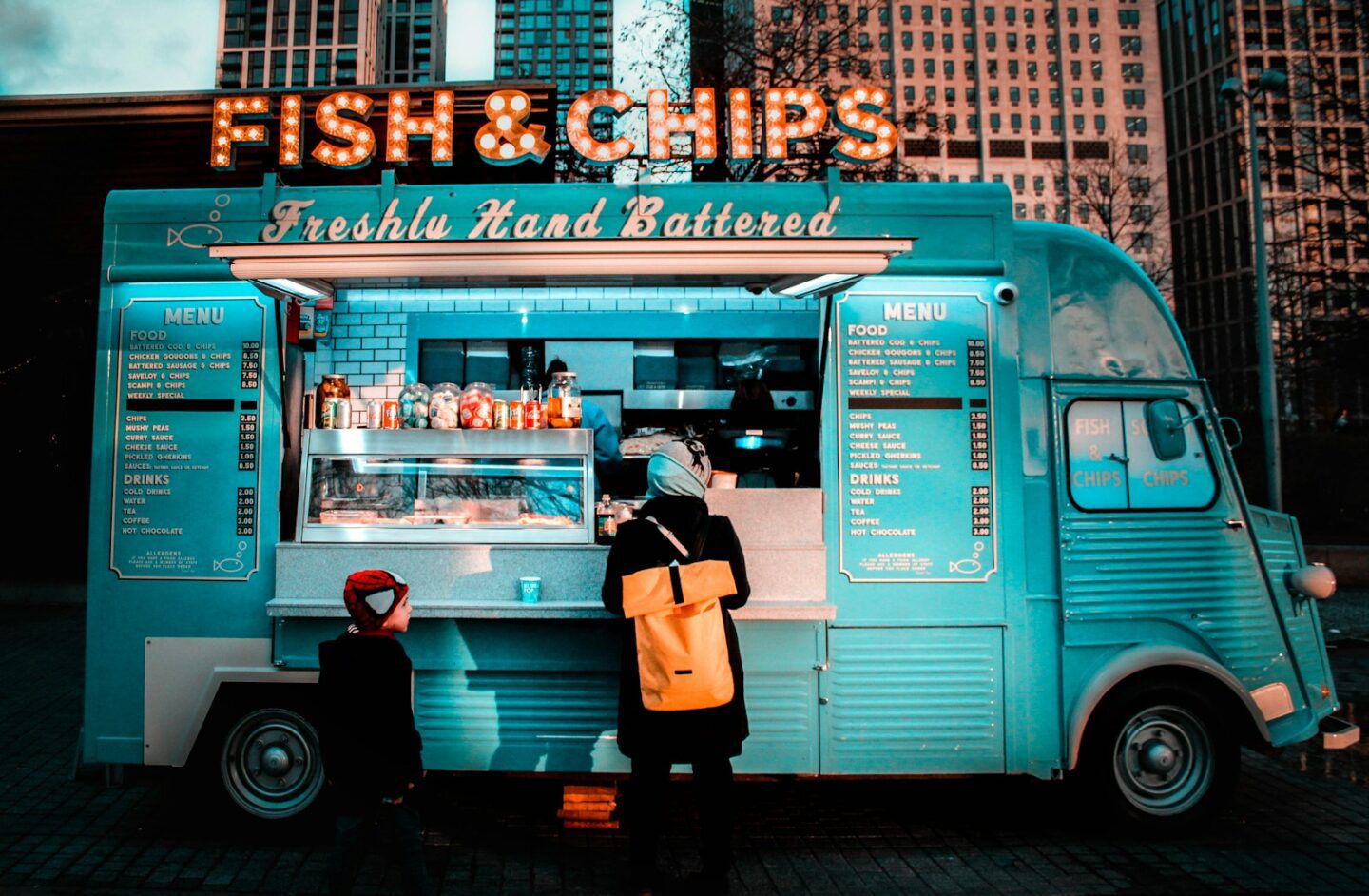 woman in brown coat standing in front of food stall with fish and chip sign
