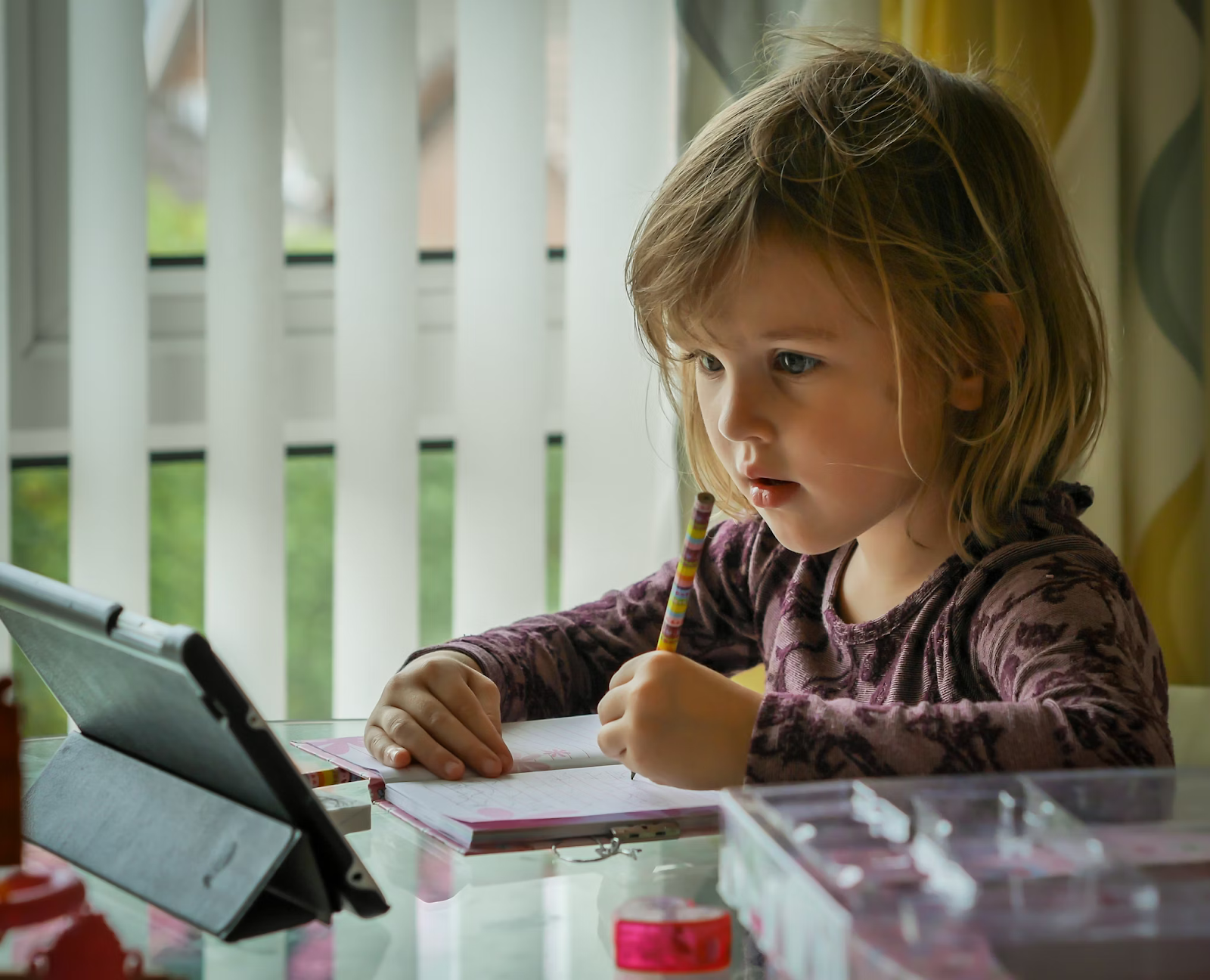 young girl sitting at table holding pen and looking at tablet