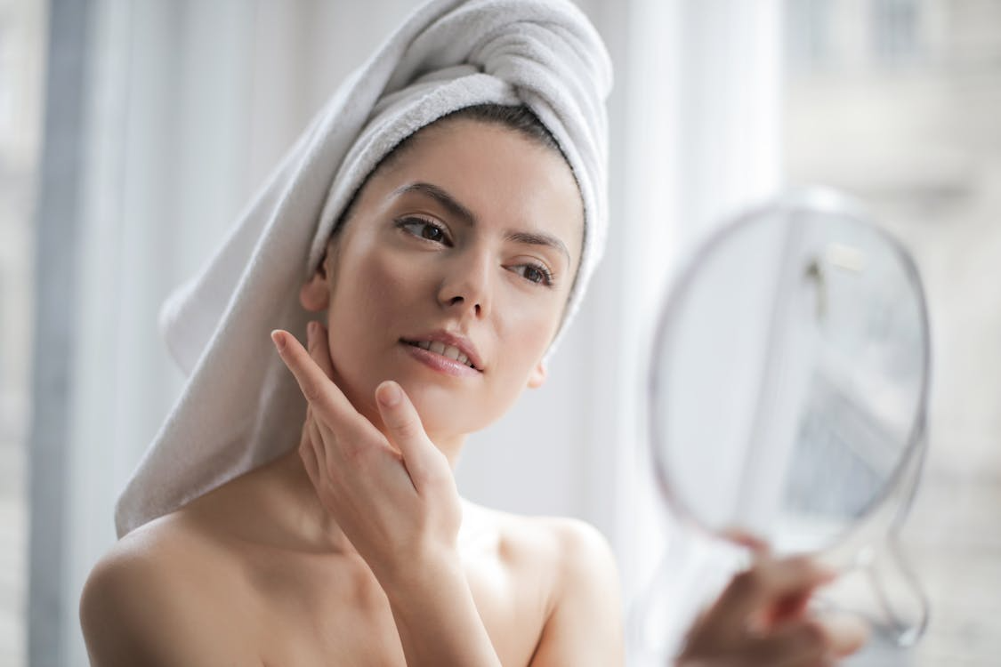woman with her hair wrapped in a towel, looking in a mirror with her hand touching her face