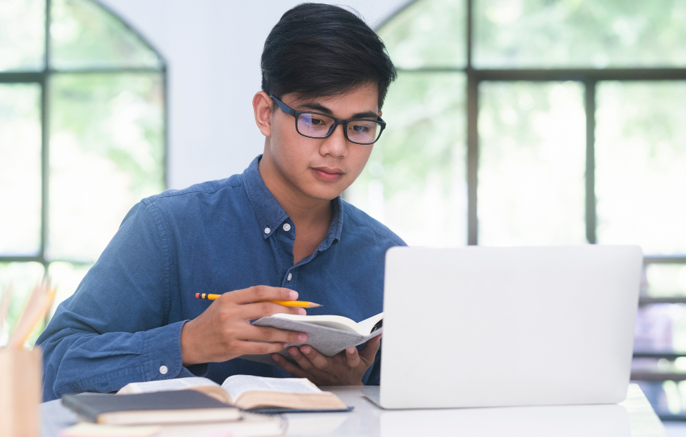 a young man sitting at desk with laptop on it, and notebook and pencil in hands