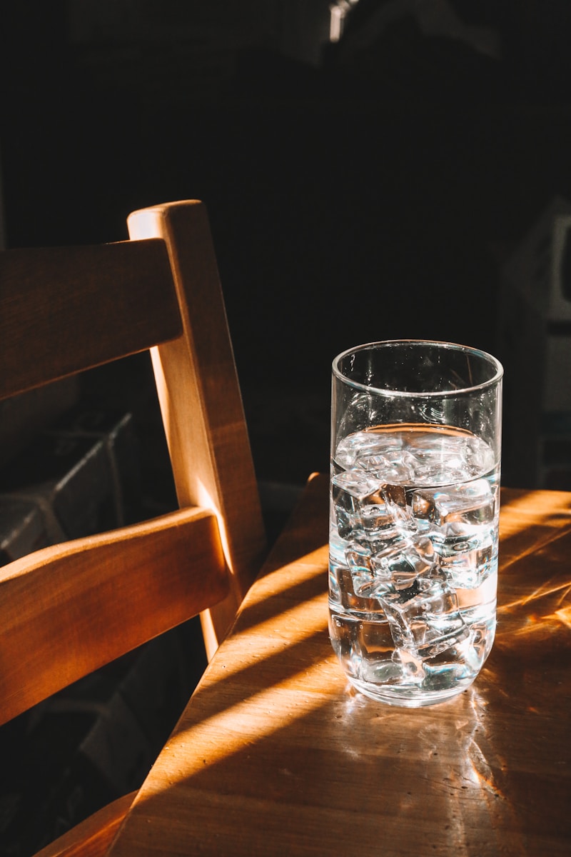 clear drinking glass full of ice and water on brown wooden table with chair next to it
